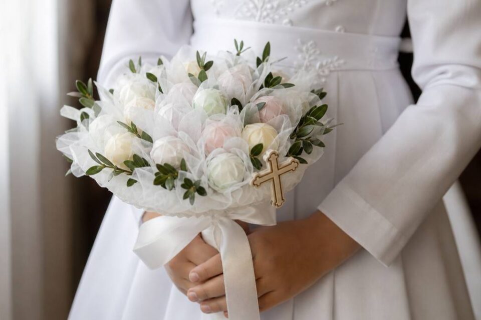 Komunijny bukiet ze słodyczy w dłoniach dziecka, z krzyżykiem wpiętym przy krawędzi kompozycji